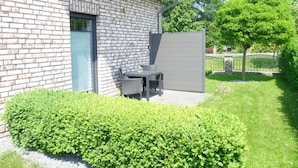 A brick building with a patio area, a door, and a large hedge in the foreground. - Holiday Home 'Melody' with Private Terrace and Wi-Fi (Ralswiek)