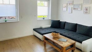 A modern living room with a sectional sofa, a wooden coffee table, and two windows with blinds. - Holiday Home 'Melody' with Private Terrace and Wi-Fi (Ralswiek)