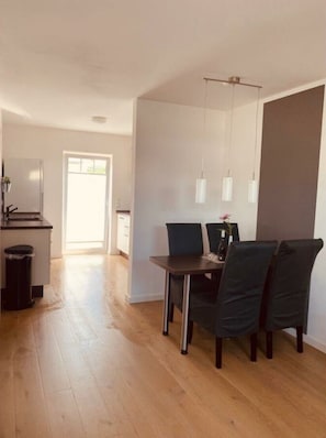A dining area with a table and chairs, a trash bin, and a kitchen in the background.