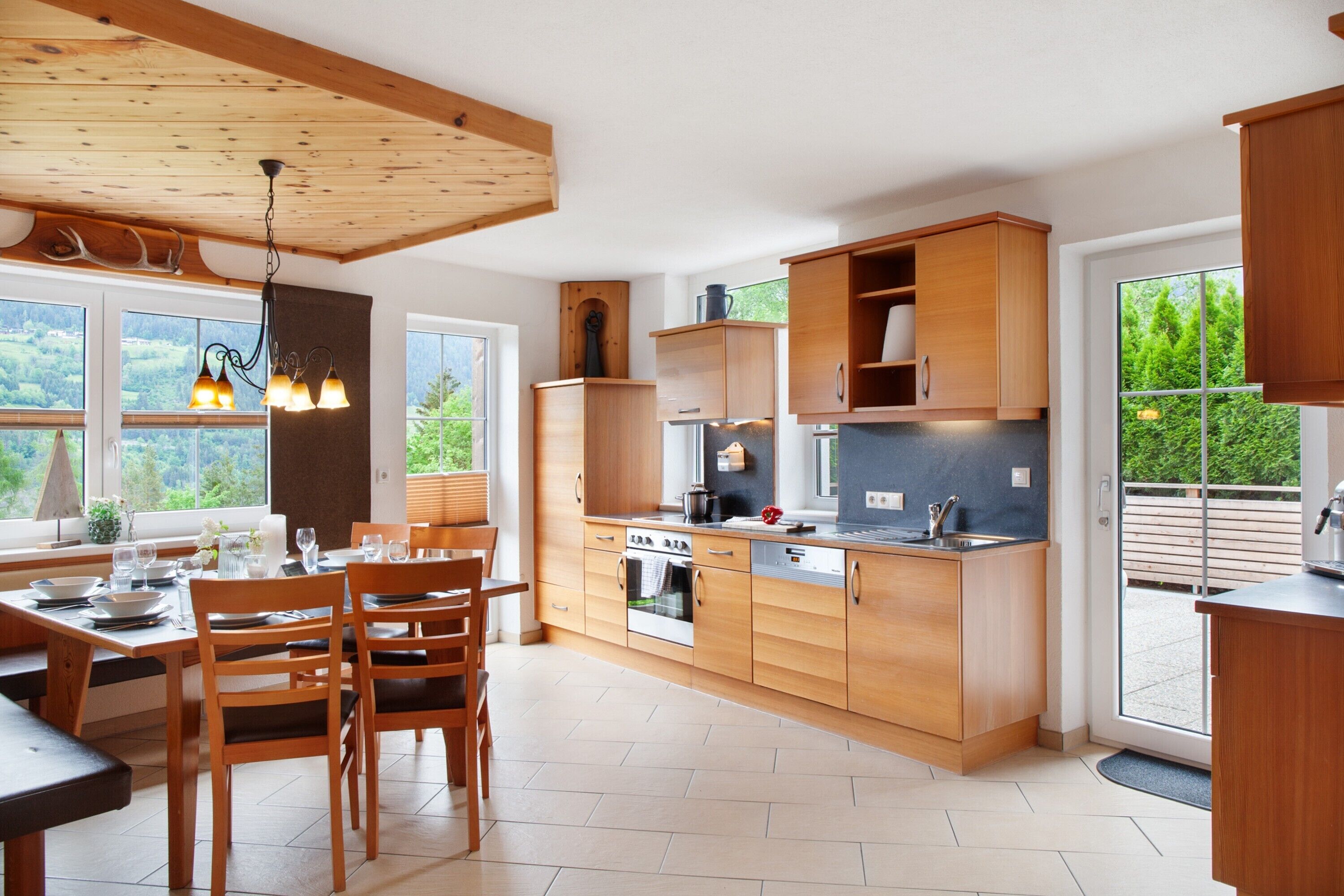 A modern kitchen with wooden cabinets, a dining area with chairs, and a large window offering a view of greenery.