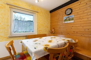Un coin repas avec une table dressée pour deux personnes, un mur en bois, une horloge et une fenêtre donnant sur un paysage.