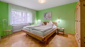 Un dormitorio con una cama de madera, un radiador, una ventana con cortinas de gasa, una mesita de noche y un armario.