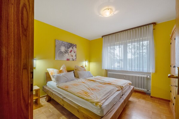 A bedroom with a wooden bed, a nightstand, a wall-mounted light, a radiator, and a window with sheer curtains.