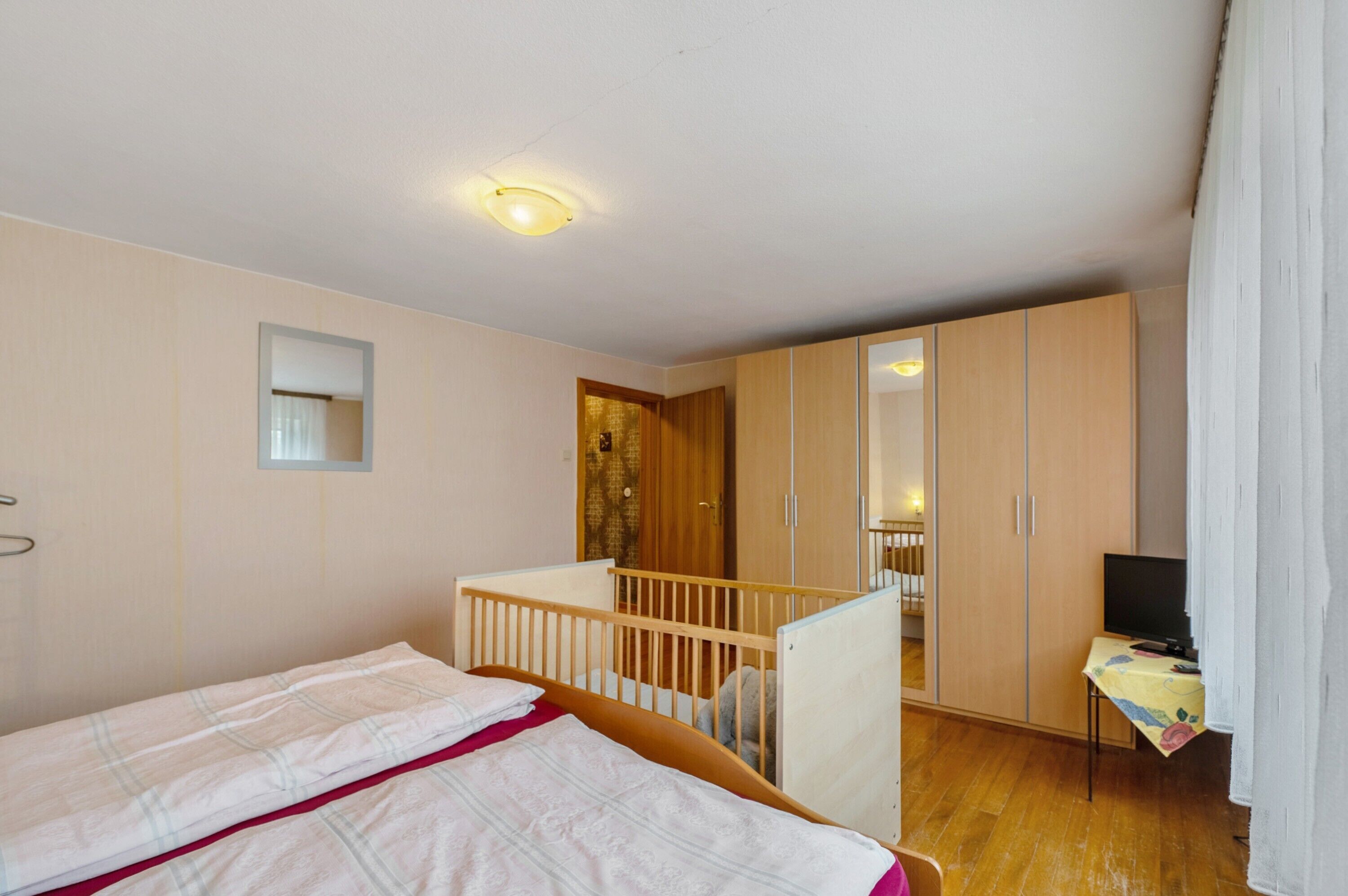A bedroom with a crib, a wardrobe, a television on a table, and a mirror on the wall.
