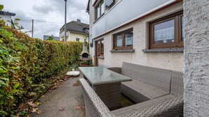 Un patio con mobiliario de mimbre, una mesa de vidrio y un seto.