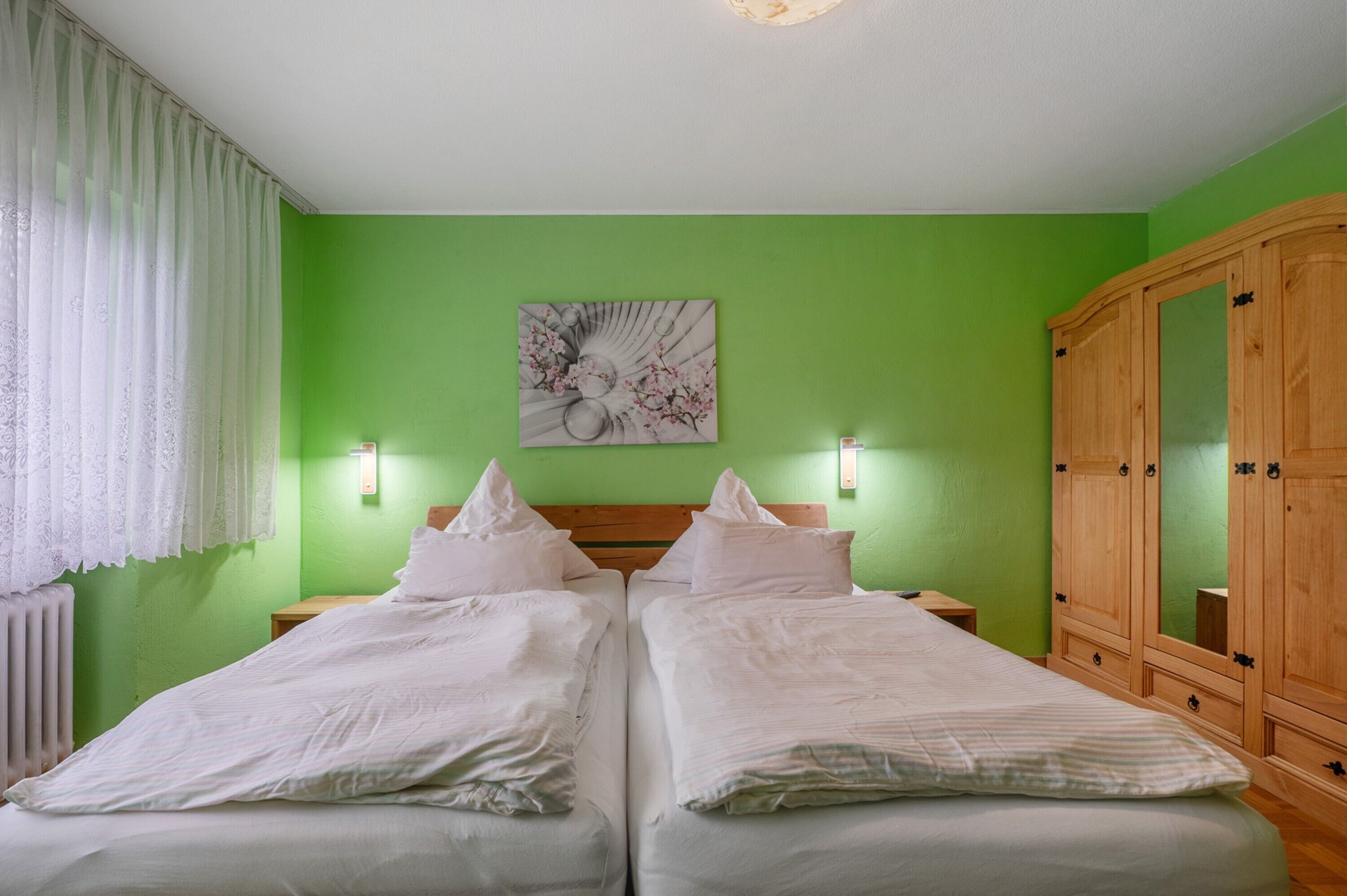 Ein Doppelbett mit weißer Bettwäsche, eine grüne Wand, ein Holzschrank und ein an der Wand befestigtes Kunstwerk.