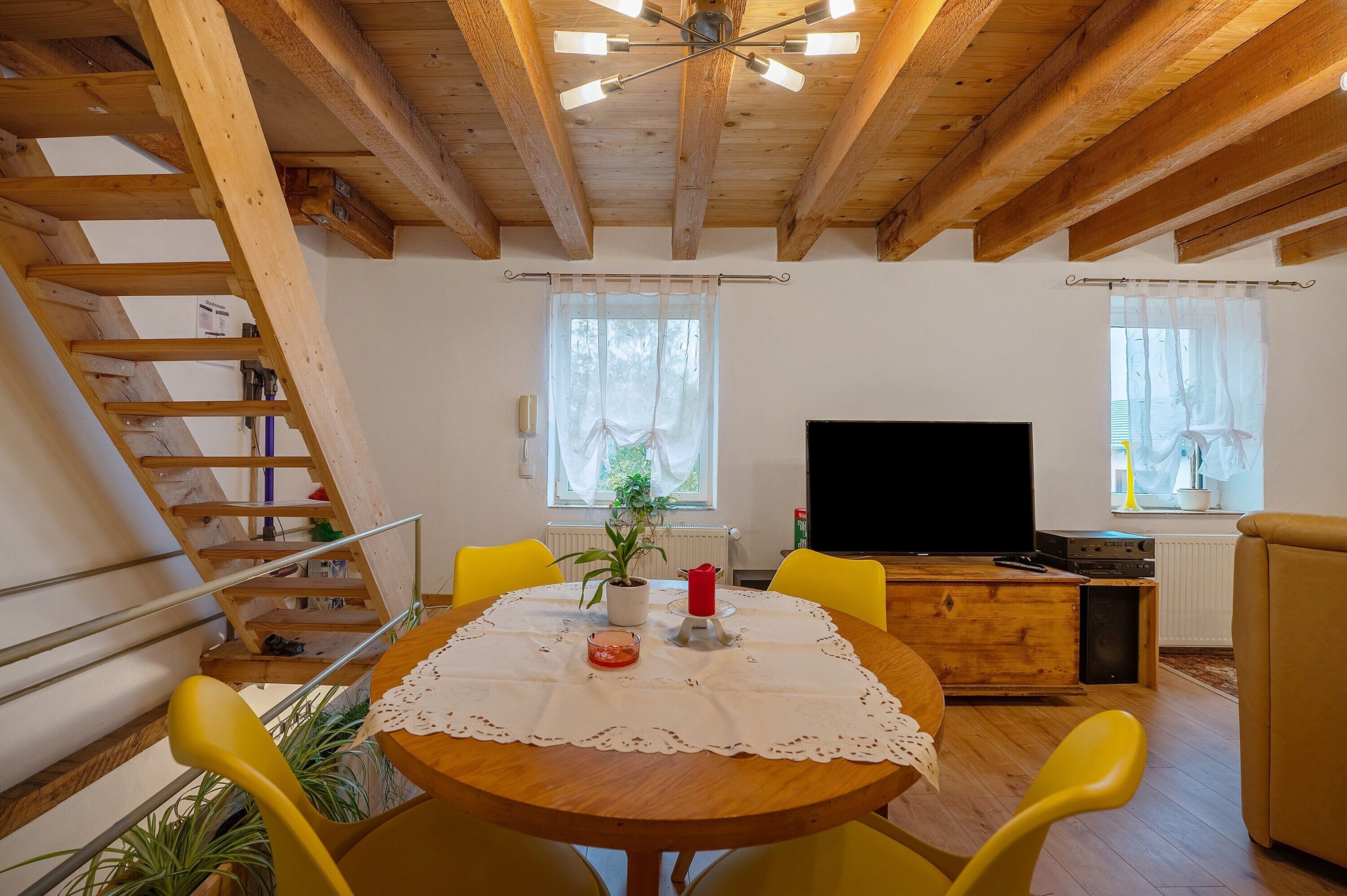 Eine Holzbalkendecke, ein Esstisch mit weißer Tischdecke, gelbe Stühle, ein Fernseher und eine Treppe.