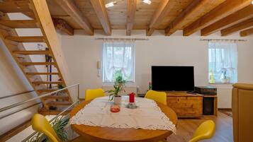 Eine Holzbalkendecke, ein Esstisch mit weißer Tischdecke, gelbe Stühle, ein Fernseher und eine Treppe.