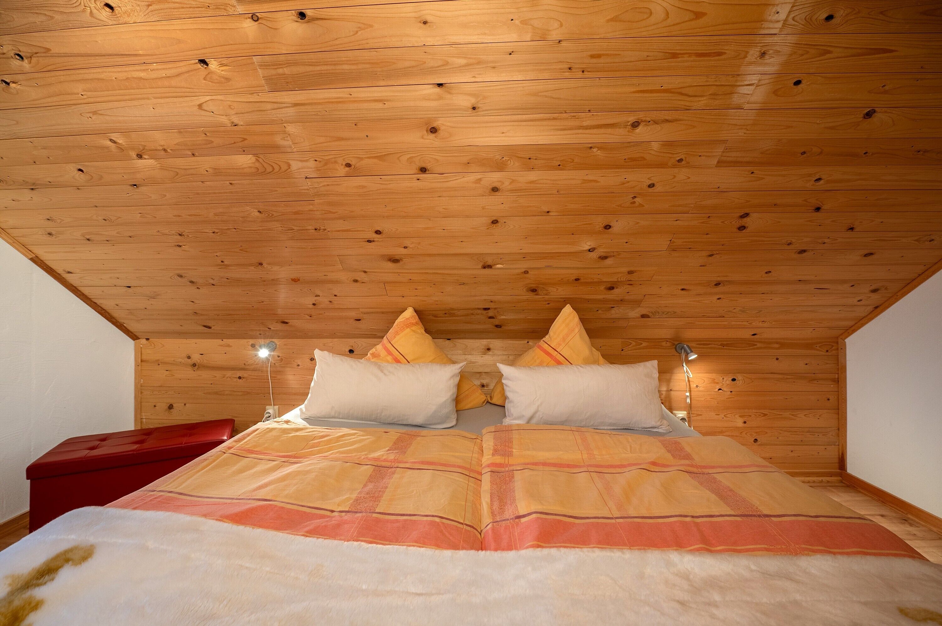 A wooden ceiling, a bed with a blanket, pillows, and a bedside lamp.