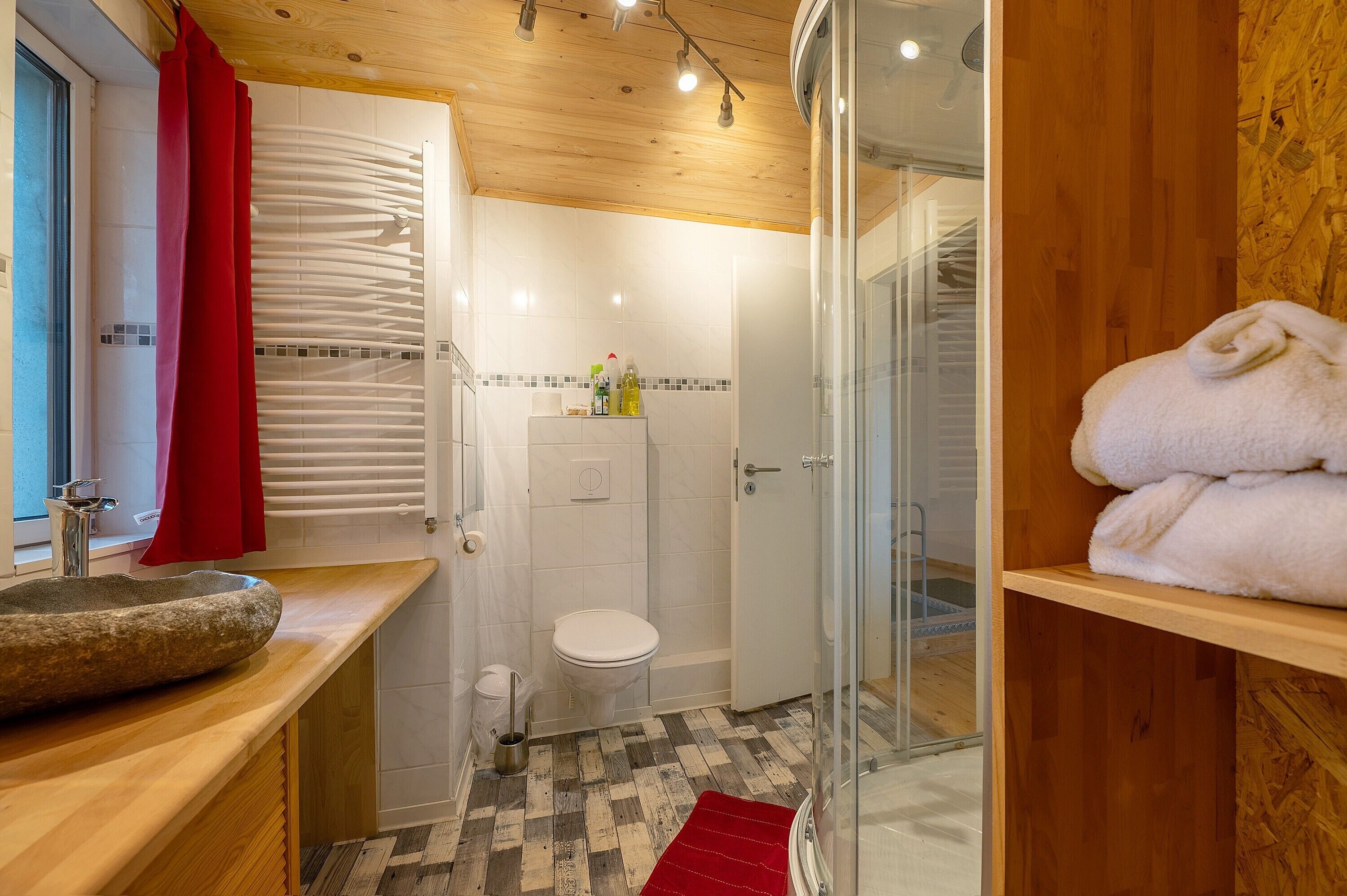A bathroom with a stone sink, white toilet, shower, and a red towel rack.