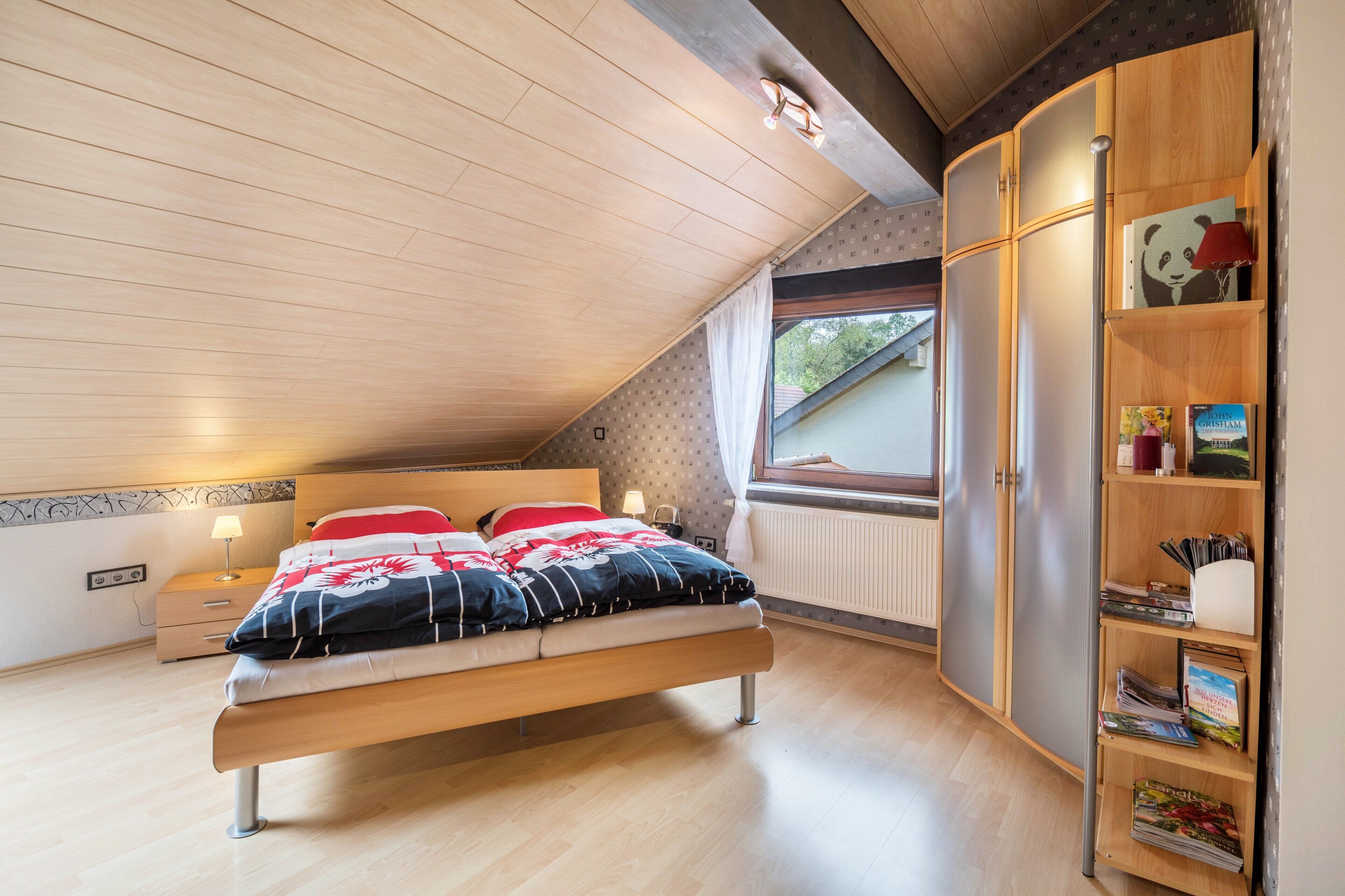 Een slaapkamer met een bed, nachtkastje, een boekenkast en een raam.