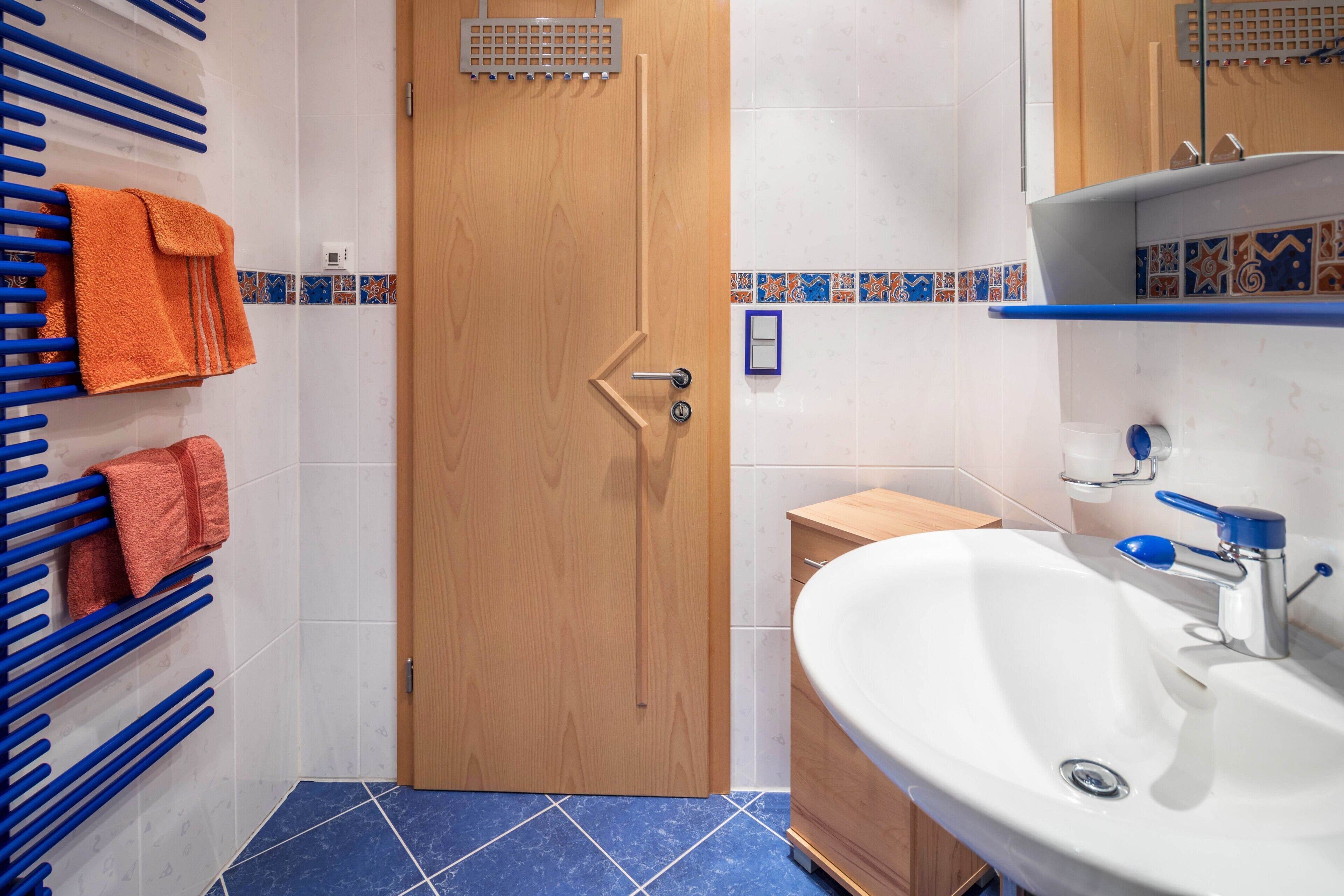 A bathroom with a wooden door, a sink with a blue faucet, and a towel rack with orange towels.