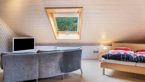 Quarto com teto inclinado, sofá, cama, televisão e claraboia.