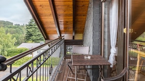 Varanda de madeira com mesa, cadeiras, grade e vista para o verde e edifícios.