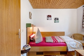 Quarto com cabeceira de madeira, cama com várias almofadas, uma mesinha e uma cadeira.