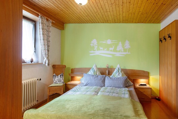 A bedroom with a bed, nightstands, a chair, a radiator, and a wall with a nature scene. - Haus Kurparkblick – Wehratal Apartment with Private Terrace and Wi-Fi (Todtmoos)