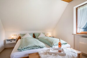Une chambre avec un plafond en pente, un lit avec une literie verte, une table de chevet avec une bouteille et un verre, et une fenêtre avec un rideau.