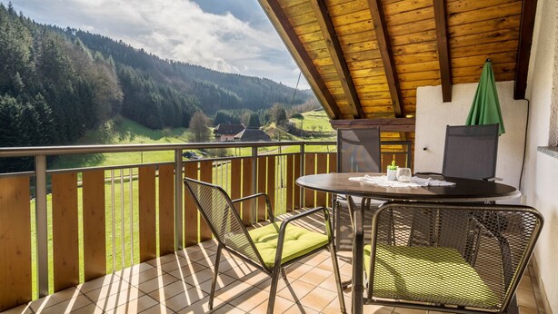 A balcony with a view of a forested valley, a table with chairs, and a green umbrella. - Apartment Morgensonne, 55 sqm, 2 bedrooms, max. 4 persons (Hofstetten)