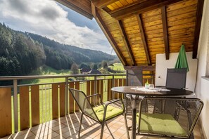 Un balcon donnant sur une vallée boisée, une table avec des chaises et un parasol vert.