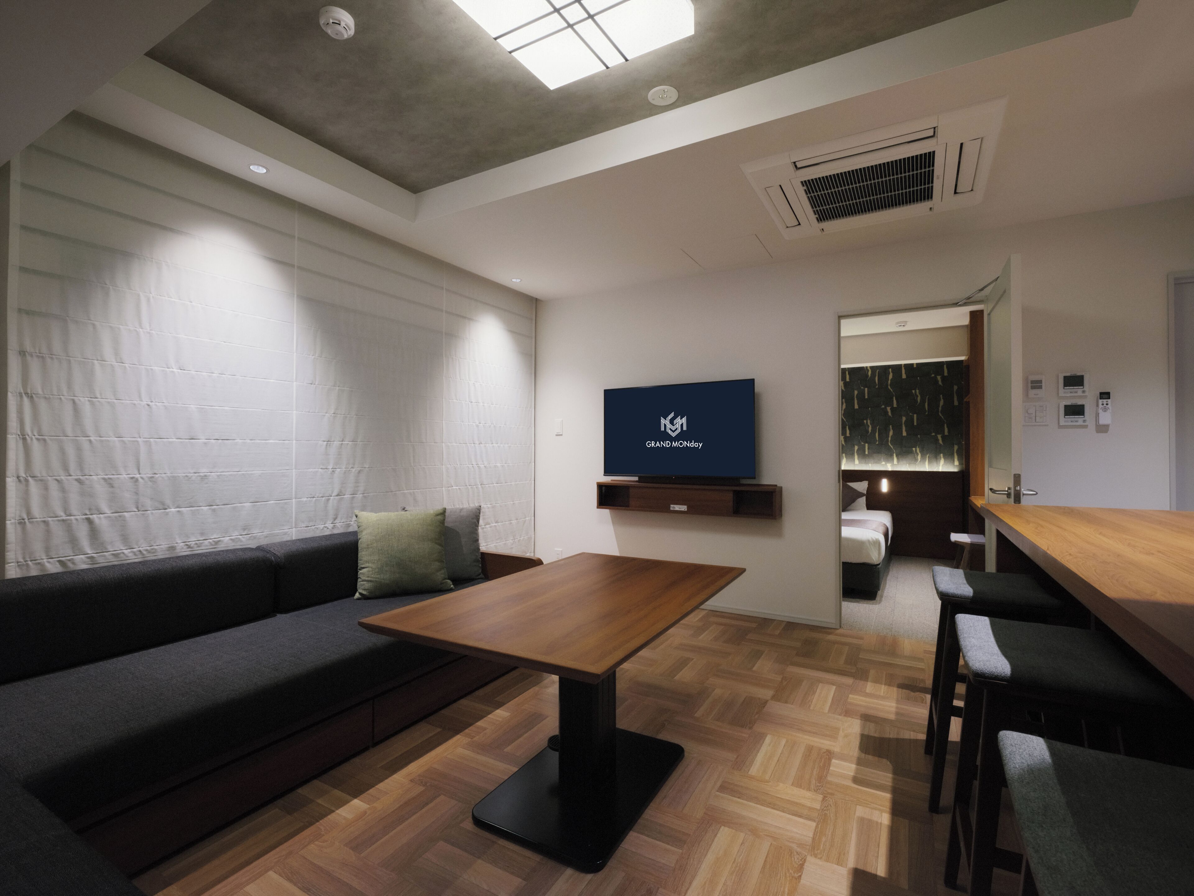 A modern hotel room with a sofa, a wooden table, a TV mounted on the wall, and a visible bathroom door.