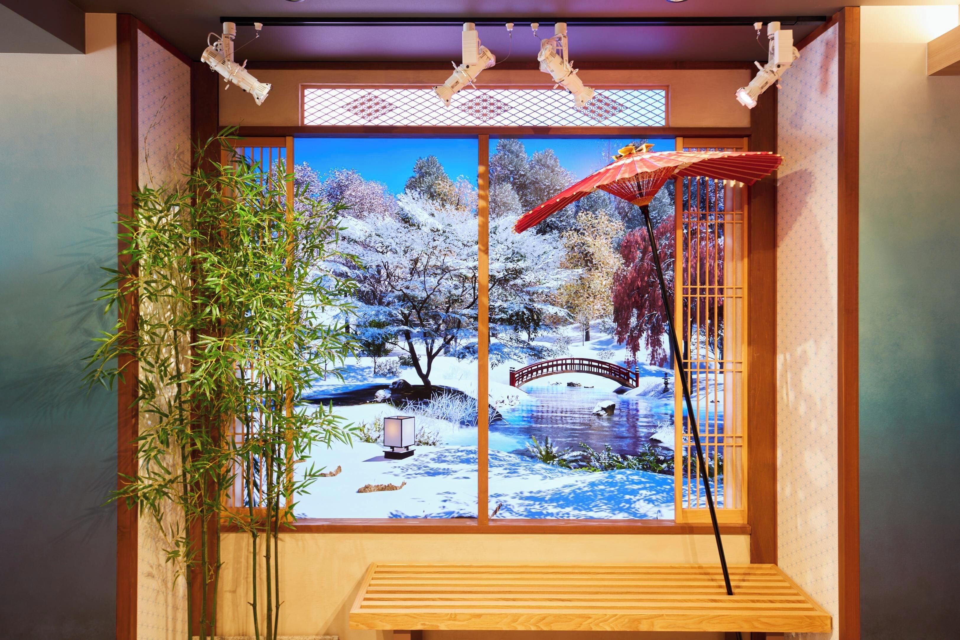 A traditional Japanese room with tatami flooring, a wooden bench, a bamboo plant, and a large window with a scenic view of a snowy landscape and a bridge.