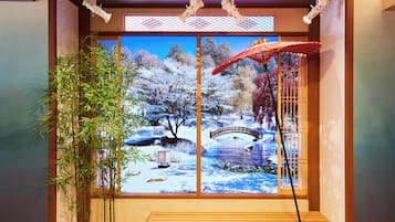A traditional Japanese room with tatami flooring, a wooden bench, a bamboo plant, and a large window with a scenic view of a snowy landscape and a bridge.
