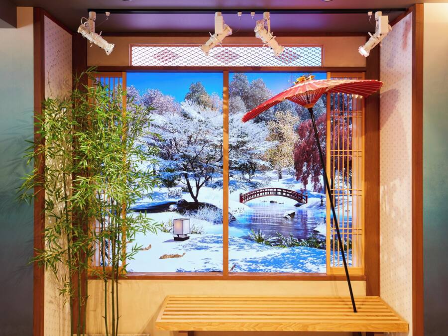 A traditional Japanese room with tatami flooring, a wooden bench, a bamboo plant, and a large window with a scenic view of a snowy landscape and a bridge.