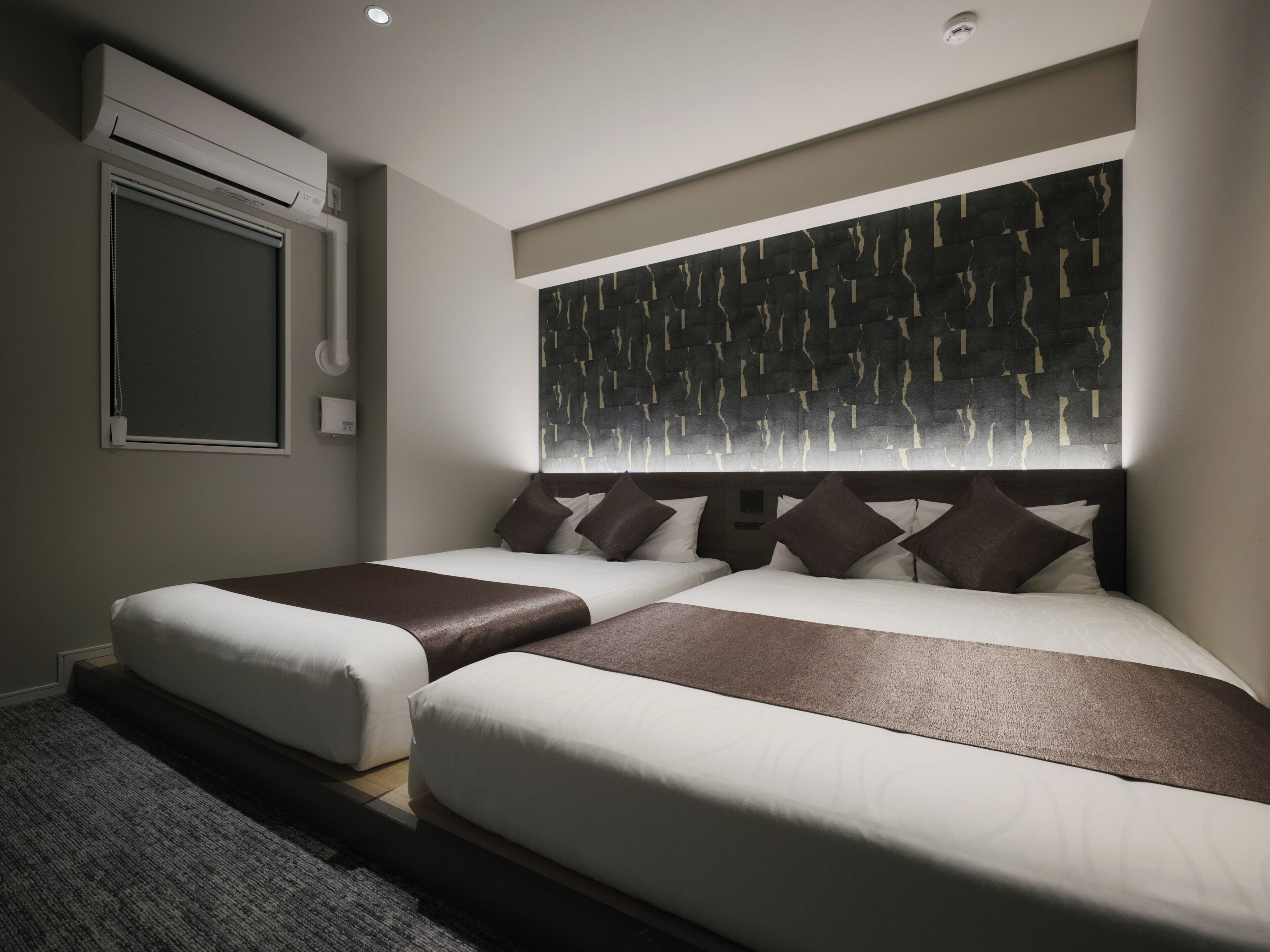 A hotel room with two beds, a large abstract wall art piece above the beds, an air conditioning unit mounted on the wall, and a window with a closed blind.