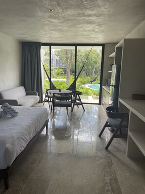 A modern hotel room with a bed, a seating area with a table and chairs, a large window with a view of greenery, and a built-in kitchen area. - Tulum Sweet condo with private pool.  Enjoy nature in the Maya jungle! (Tulum)