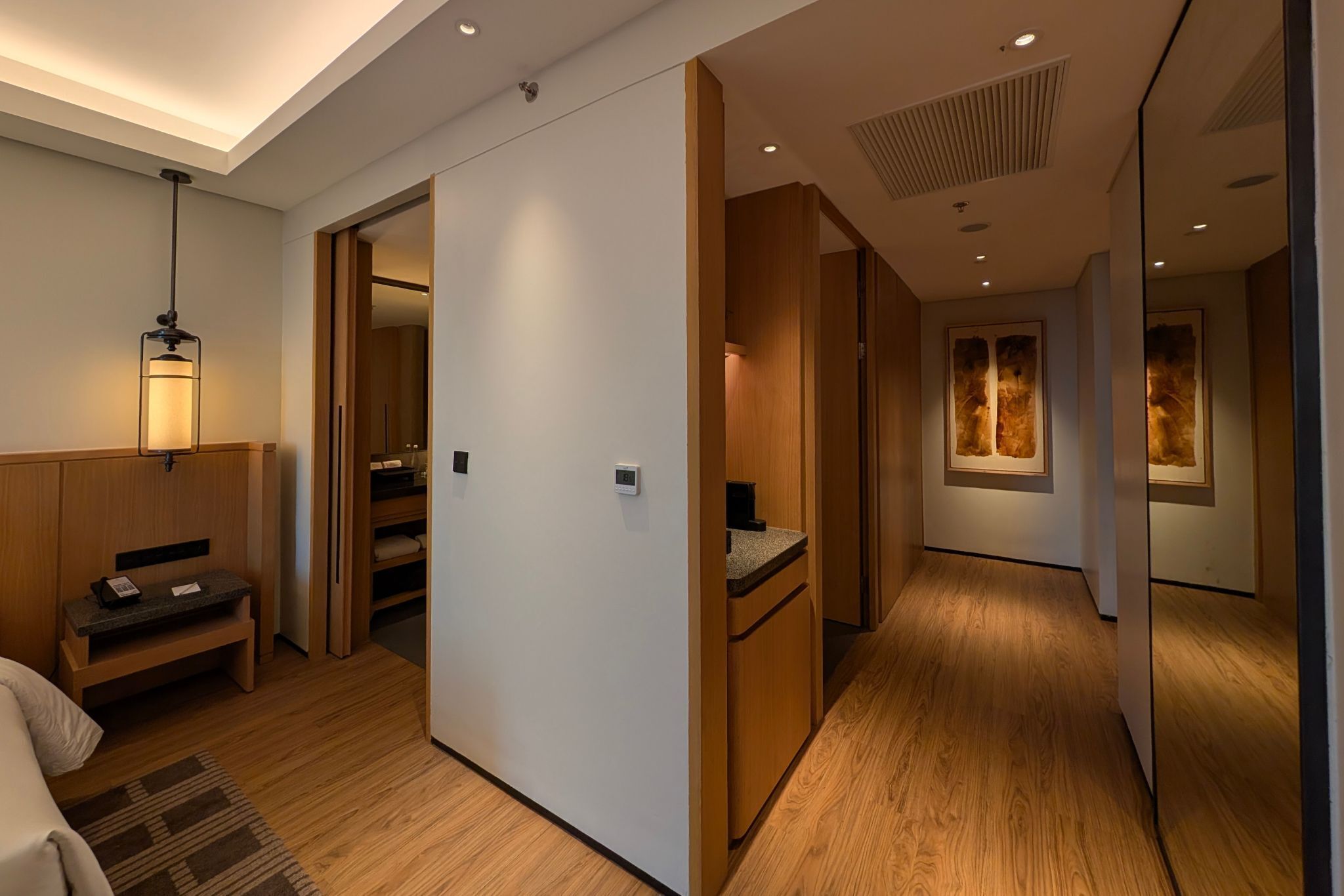 Habitación de hotel con piso de madera, un escritorio con teléfono, un banco y una lámpara fijada a la pared.