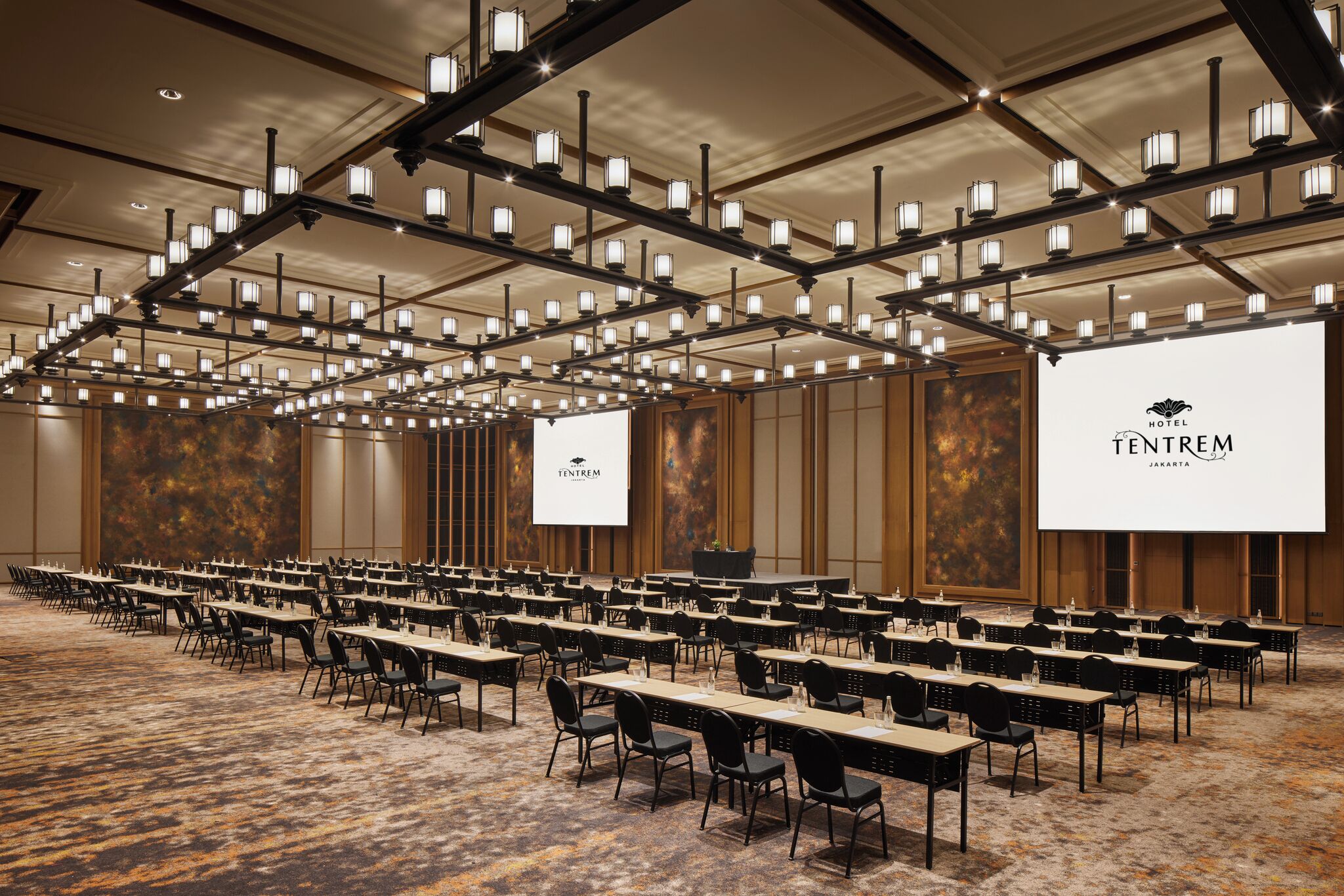 A large conference room with rows of tables and chairs, a high ceiling with hanging lights, and a large screen displaying the hotel name.