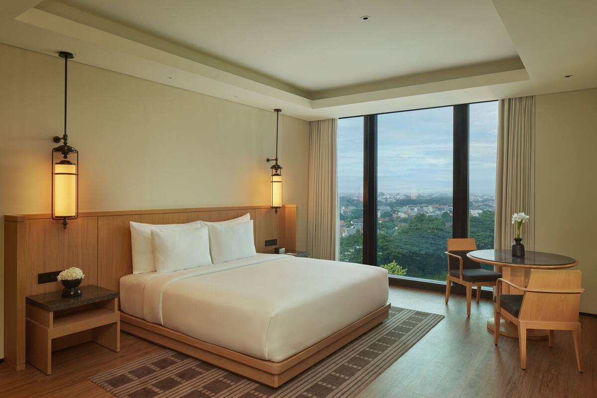 A hotel room with a large bed, two bedside tables, a round table with chairs, and a view of a cityscape through large windows.