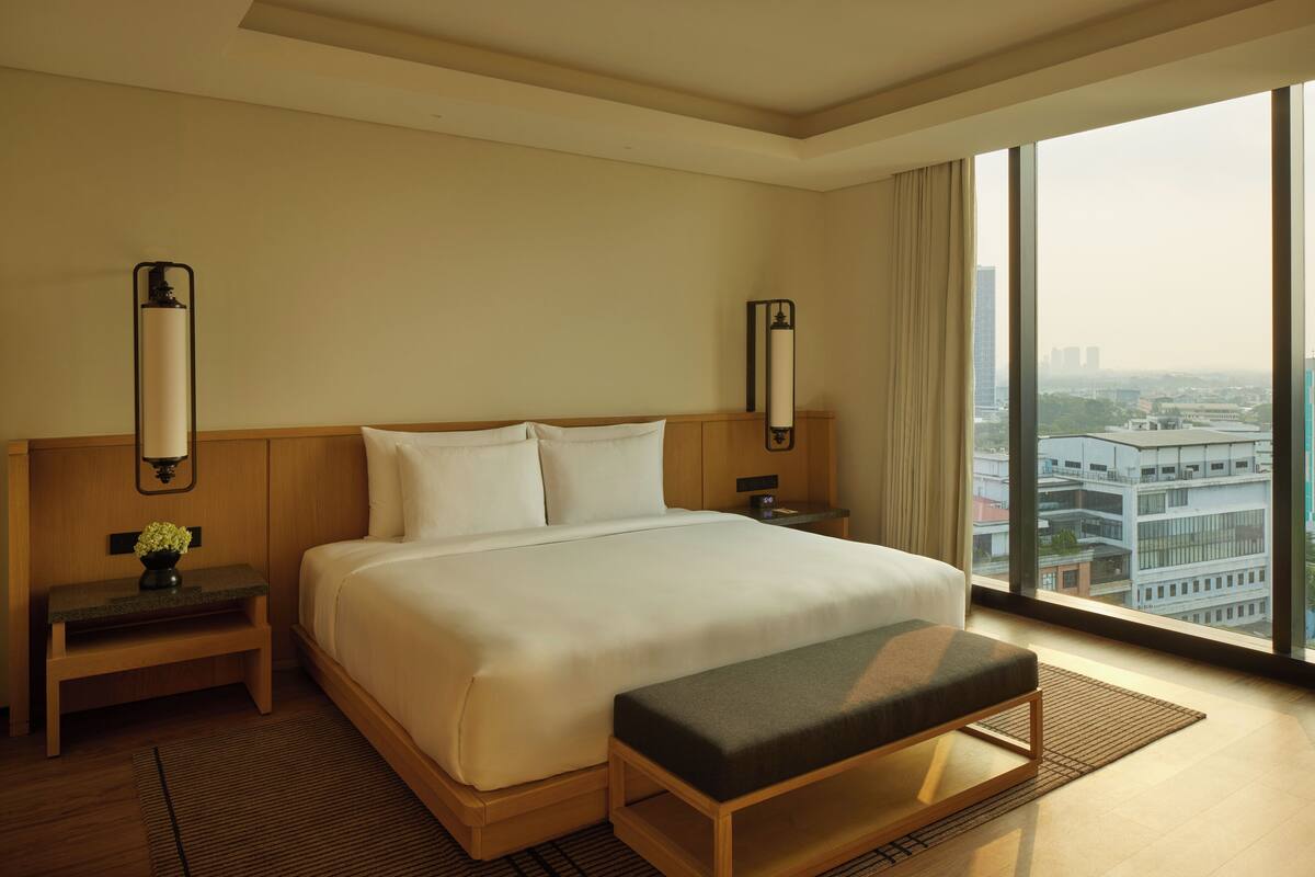 A hotel room with a large bed, a bench, a nightstand with a lamp, and a view of the city through large windows.