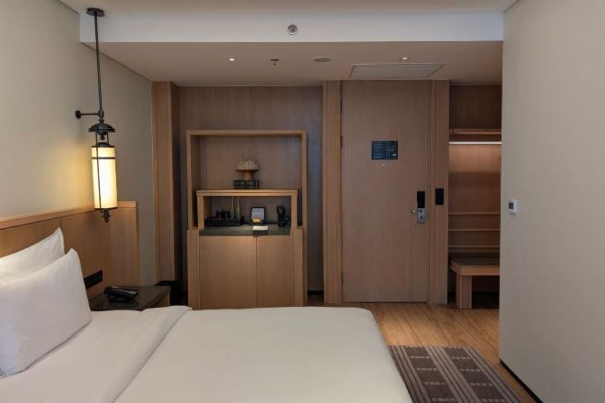 A hotel room with a bed, bedside table, lamp, and a built-in desk with a lamp.
