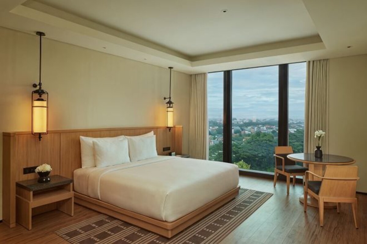 A hotel room with a large bed, bedside lamps, a small table with chairs, and a view of the city.
