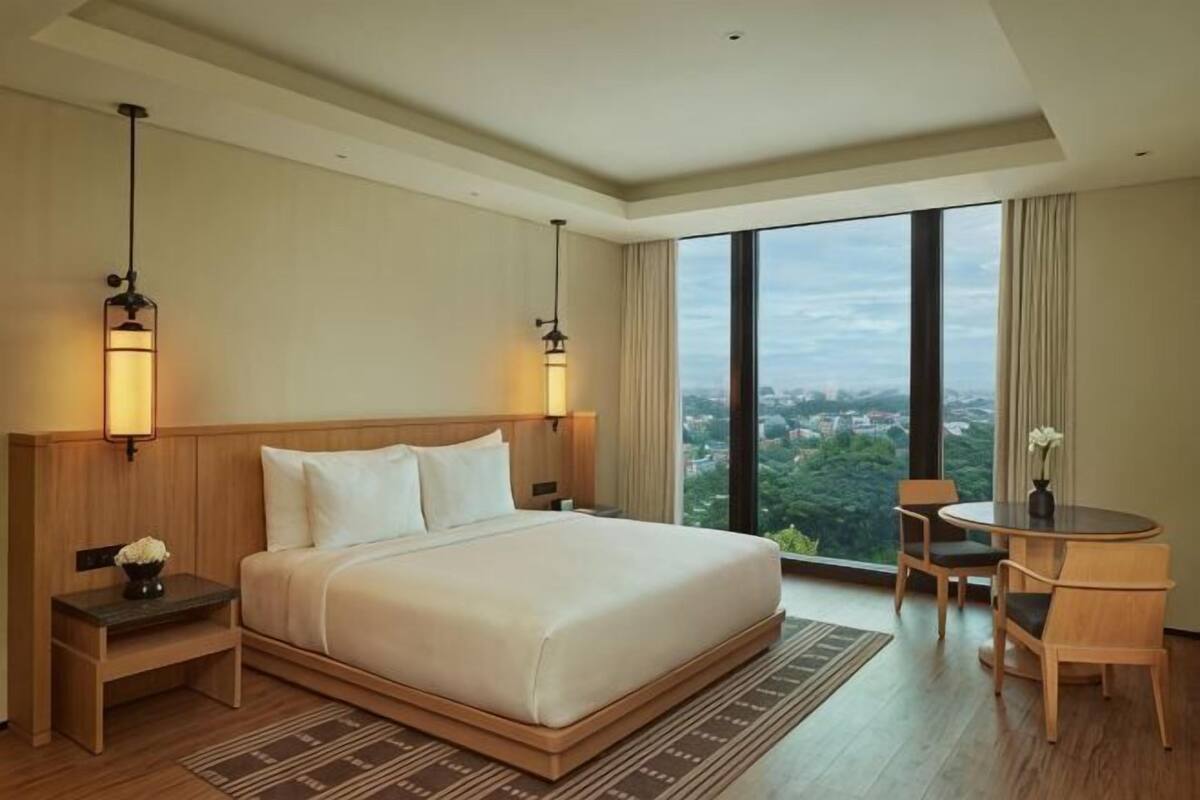 A hotel room with a large bed, bedside lamps, a small table with chairs, and a view of the city.