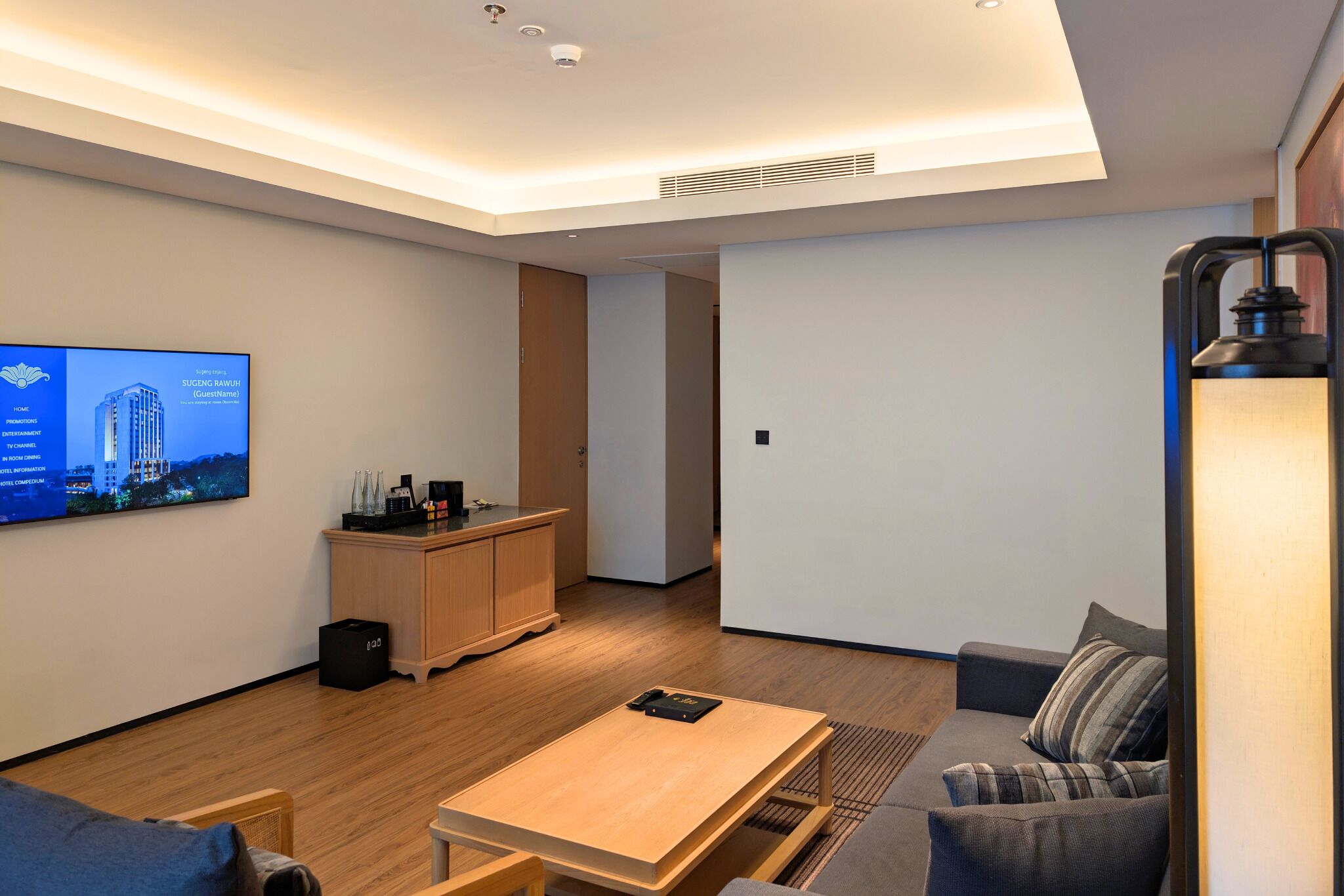 Une chambre d’hôtel moderne avec un plancher en bois, un téléviseur à écran plat fixé au mur, une petite table en bois, un canapé et une lampe.