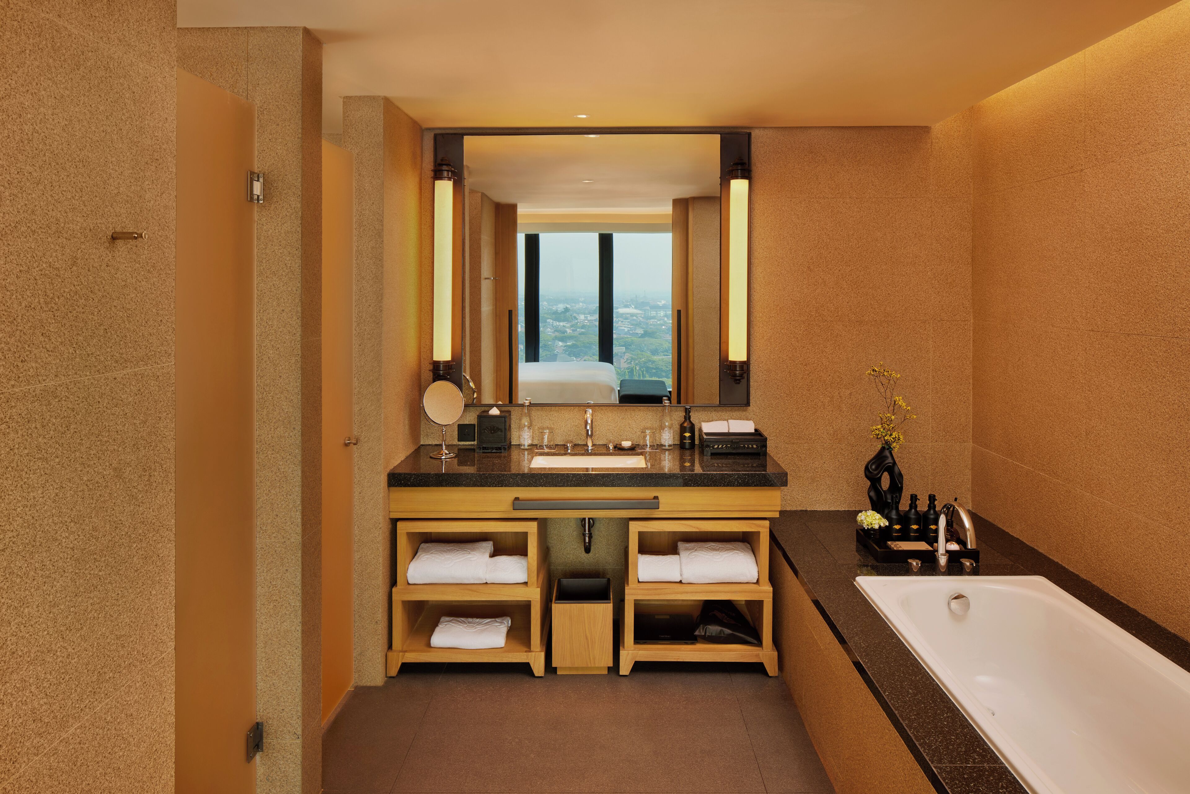 A modern bathroom with a large sink, mirror, and bathtub. It features wooden cabinetry and a view through the window.