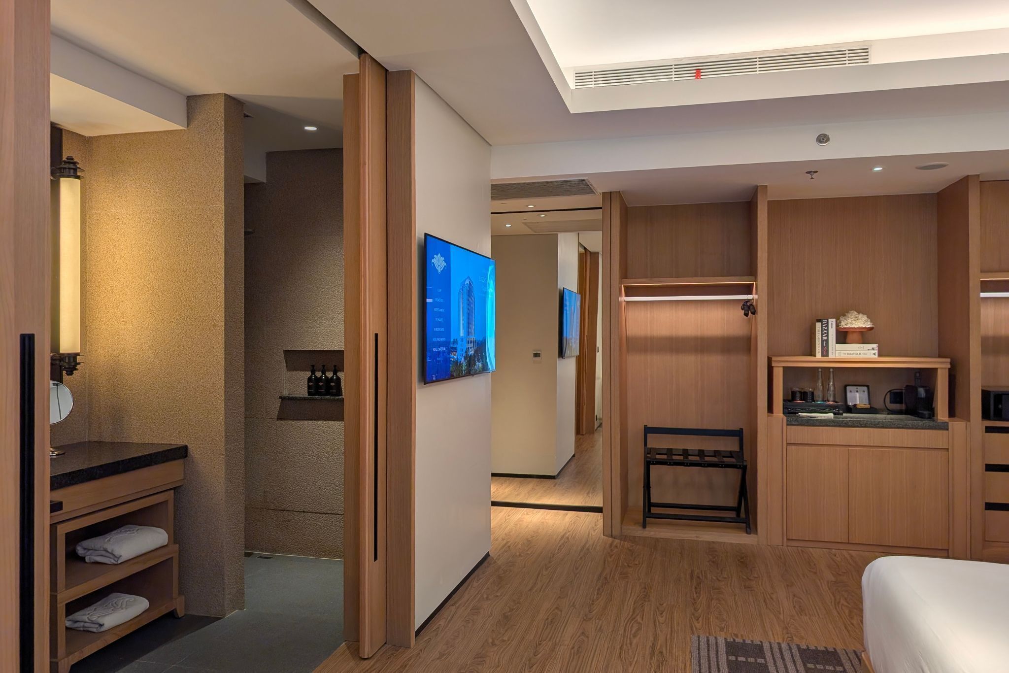 Een hotelkamer met houten vloer, een bed, bureau, stoel, kledingkast en een badkamer met wastafel en spiegel.