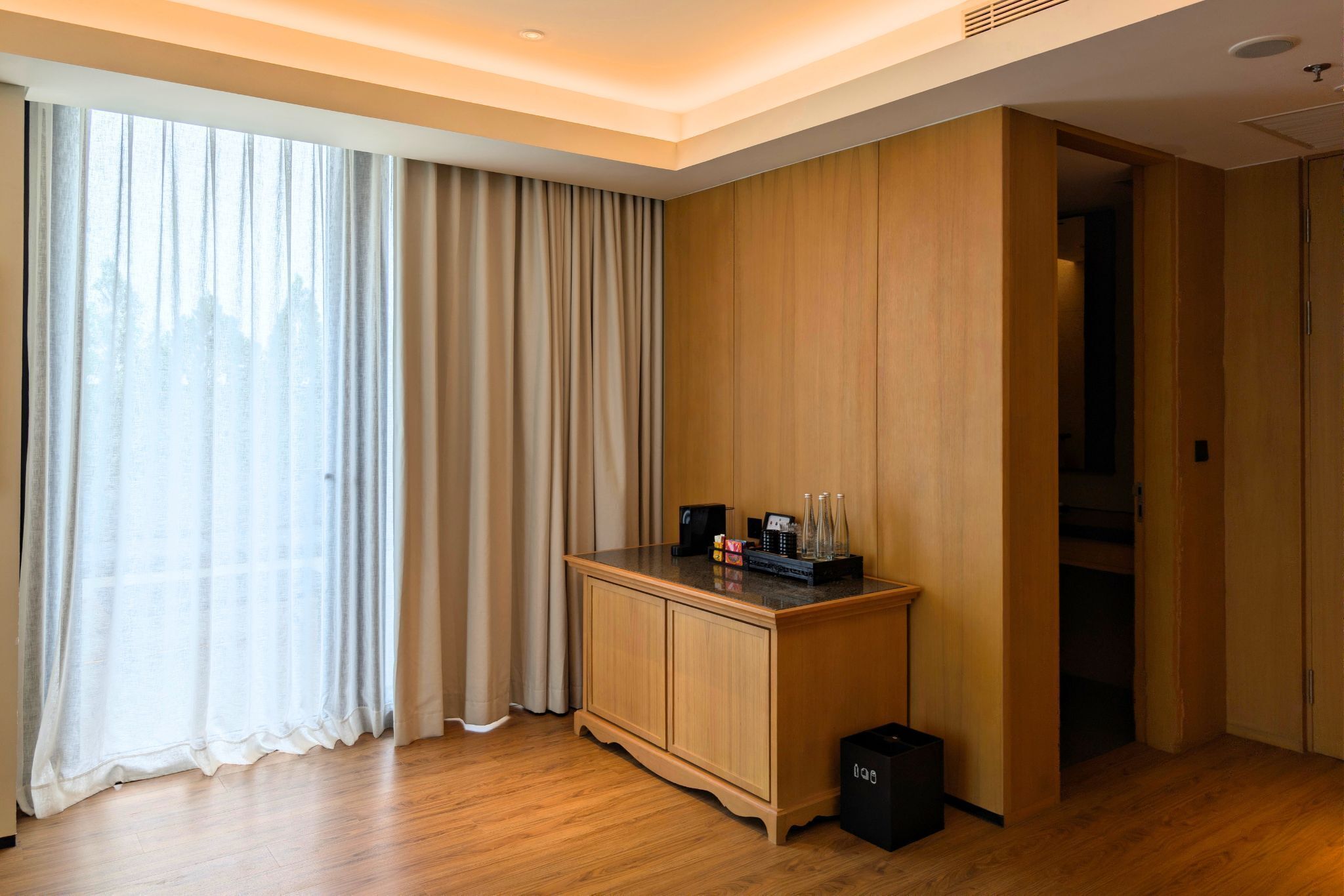 A hotel room with a wooden cabinet, a small table with a clock and bottles, and a large window with curtains.
