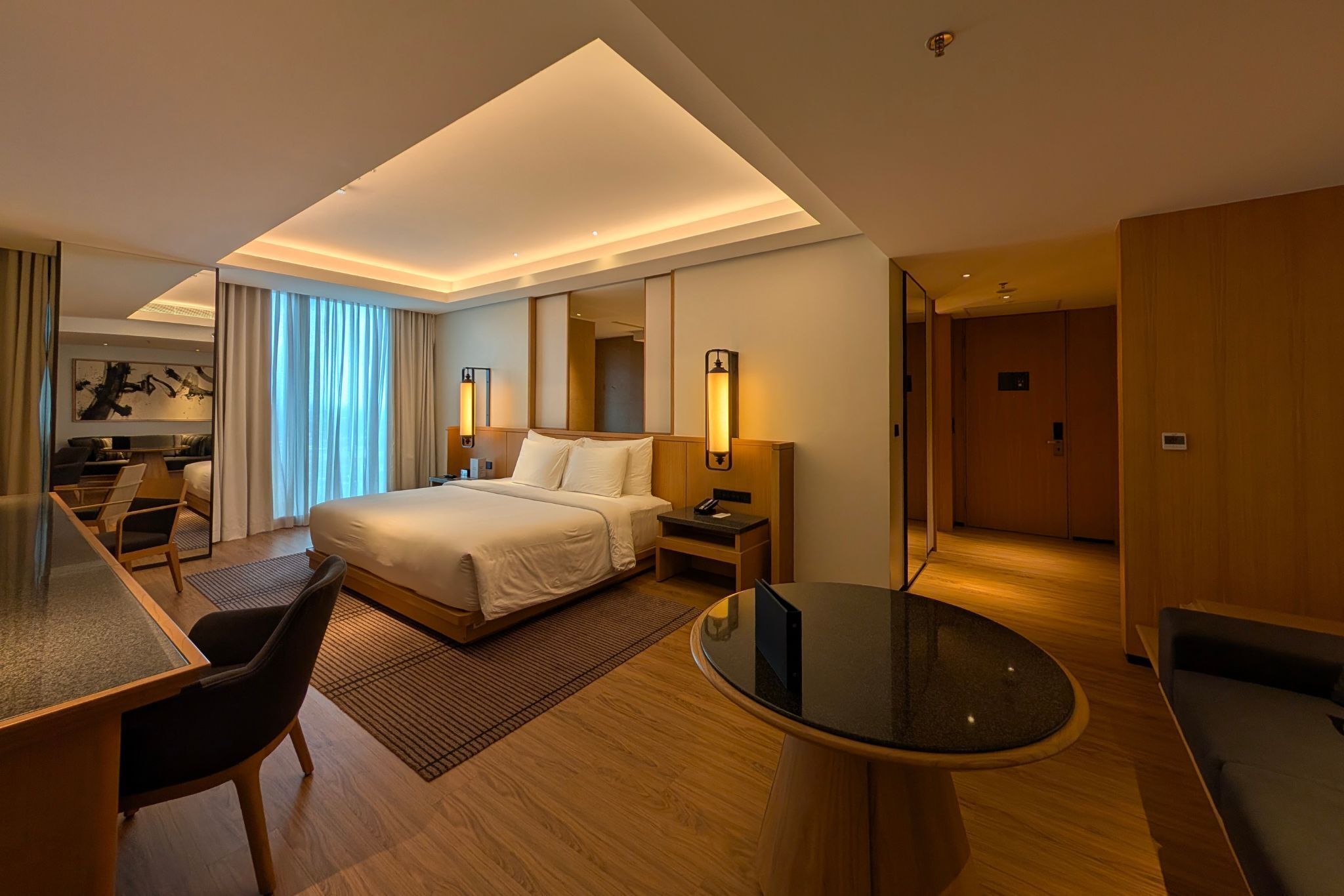 A modern hotel room with a large bed, a desk, a chair, a round table, and a separate seating area.