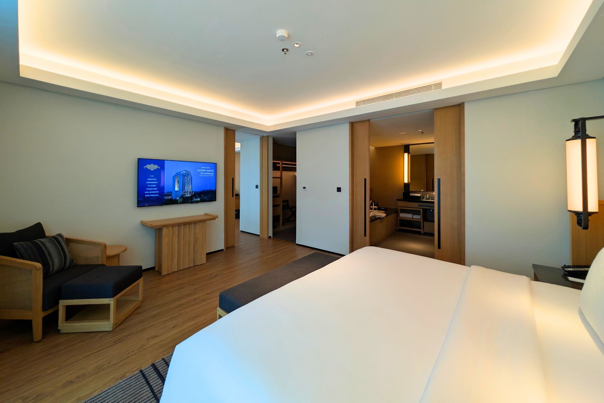 Ein modernes Hotelzimmer mit einem großen Bett, einem an der Wand montierten Fernseher, einer Sitzecke mit kleinem Tisch und einem begehbaren Kleiderschrank.