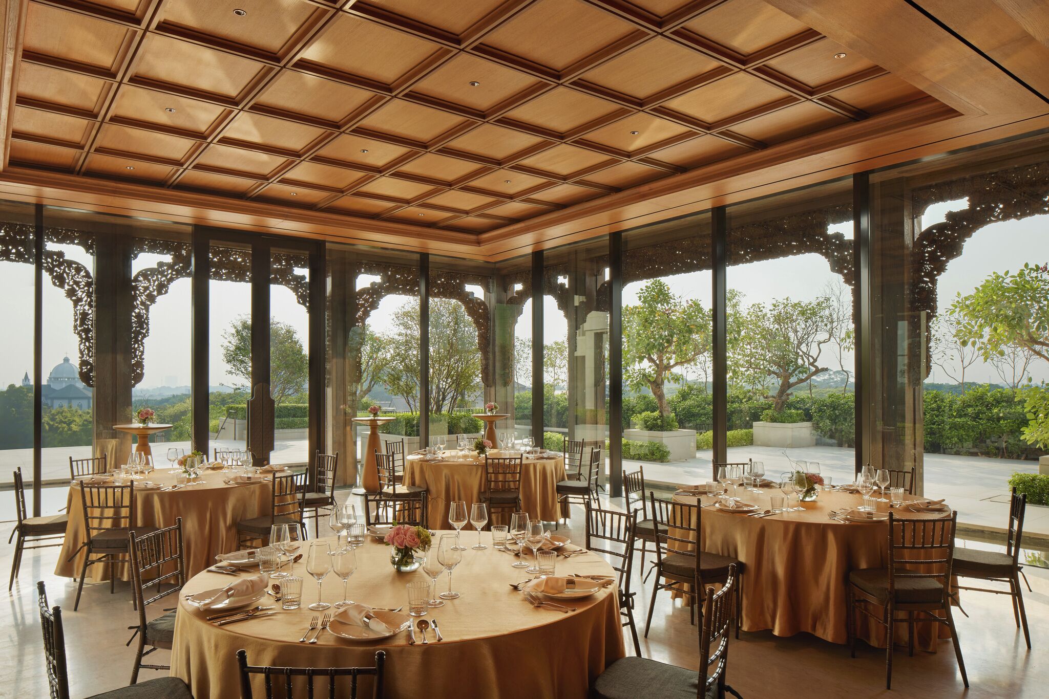 A restaurant with round tables set for dining, large windows, and a view of greenery and a building in the distance.