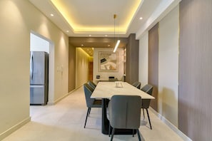 A modern dining area with a long table, chairs, recessed ceiling lighting, and a refrigerator in the background.