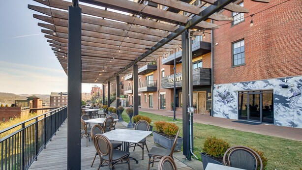 Un patio al aire libre con mesas y sillas de madera, un edificio de ladrillo con un mural y vistas de la ciudad.