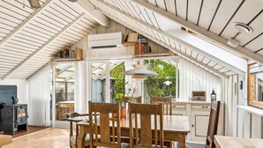 A cozy, open-plan living space with a high ceiling and wooden beams, featuring a dining area with a table and chairs, a kitchen area with a radio, and a view of the garden through large windows.