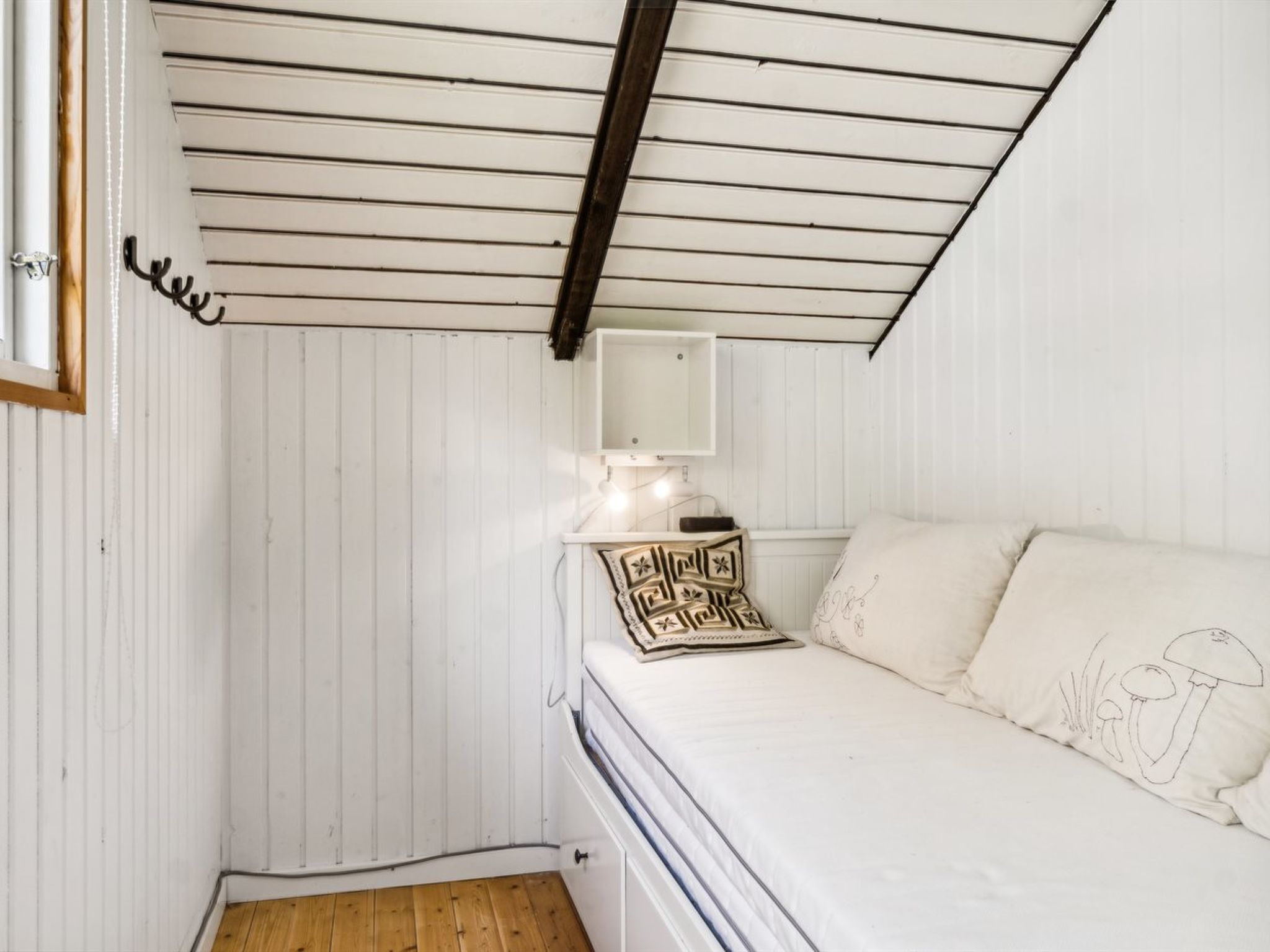 Ein kleiner Raum mit weißen Wandverkleidungen und schrägen Wänden, ein Bett mit weißer Bettwäsche, ein Nachttisch mit Lampe und ein an der Wand befestigtes Regal.