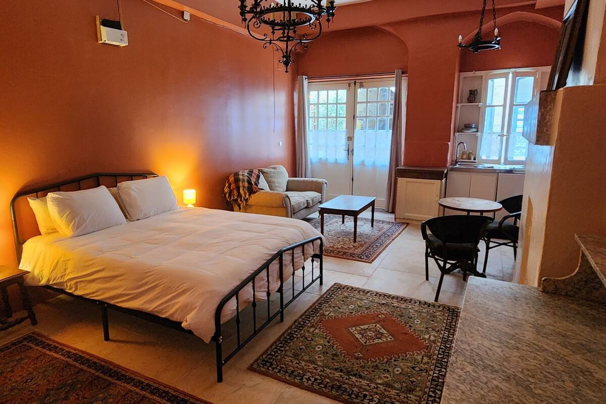 Une chambre à coucher comprenant un lit, des tables de chevet, une chaise, une petite table et donnant sur la cuisine.
