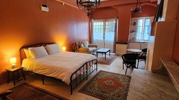 A bedroom with a bed, bedside tables, a chair, a small table, and a view of the kitchen.