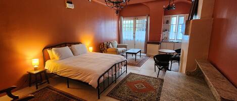 A bedroom with a bed, bedside tables, a chair, a small table, and a view of the kitchen.